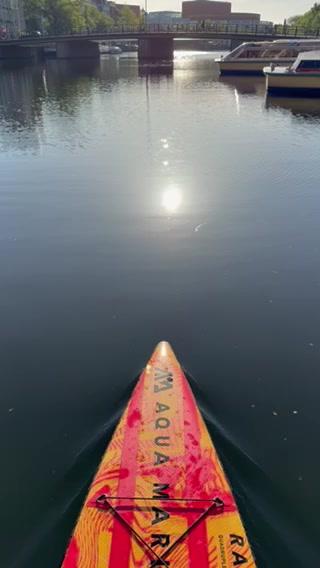 The bow of a paddle board floats on calm water with a bridge and boats in the background